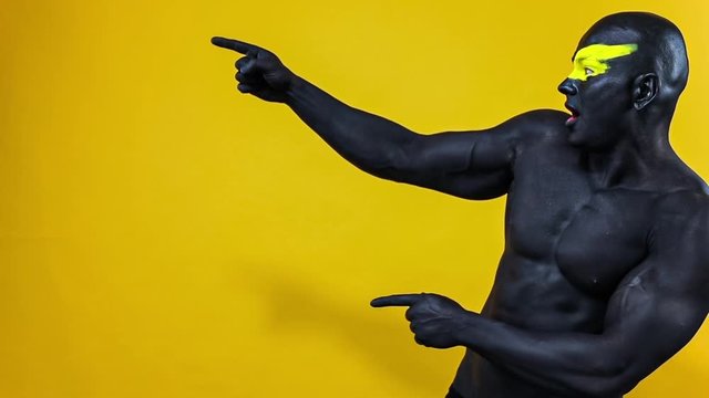 Man pick on copy space. Bodybuilder athlete with yellow face art and black body paint. Colorful portrait of the guy with bodyart. Slow motion.