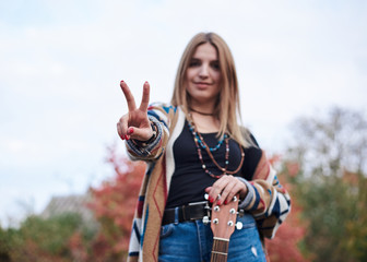 Hippie musician with acoustic guitar, showing piece sign with two fingers. Three-quarter portrait of young woman, wearing colorful cardigan and blue jeans, posing in forest park woods in autumn.
