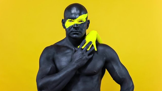 Man with female hands on the body. Bodybuilder athlete with yellow face art and black body paint. Colorful portrait of the guy with bodyart. Slow motion.