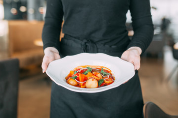 Hot dish with meat and vegetables in a white plate. The waiter carries the dish in the restaurant.