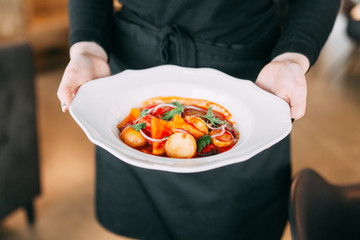Hot dish with meat and vegetables in a white plate. The waiter carries the dish in the restaurant.