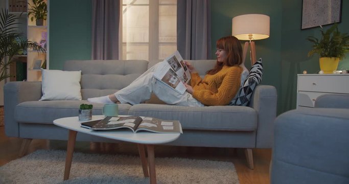 Caucasian pretty woman with red hair lying on sofa and reading magazine in cozy living room. Female homey in glasses resting in comfortable flat on weekend and flipping pages of journal lazy.