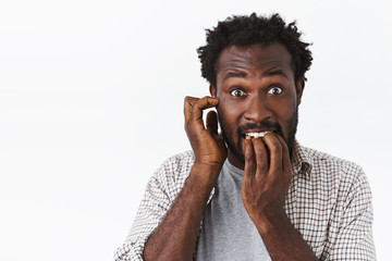 Embarrassed and worried bearded african-american guy feel panic, biting nails and scratch ear,...