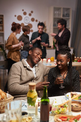 Happy African couple sitting at the table laughing and drinking wine at the party with other young people talking in the background