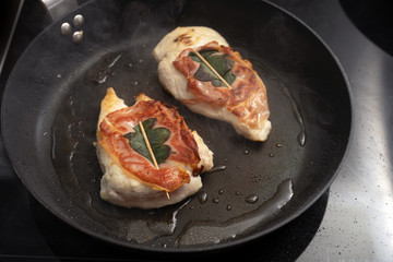 Roasted chicken saltimbocca wrapped with air-dried ham and sage leaves in a black pan on the stove, copy space