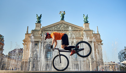 Fototapeta premium Front view of cyclist standing on front wheel of mountain bike and looking down. Young athlete is cycling downtown near fountain. Concept of active.