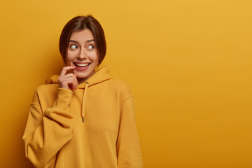 Positive good looking woman has nice idea, looks mysteriously aside, smiles gently, wears casual sweatshirt, makes up plan, has combed dark hair, isolated on yellow background, empty space right