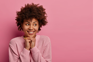 Fototapeta na wymiar Studio shot of attractive ethnic curly woman looks gladfully aside, keeps hands under chin, has toothy smile, stands in lovely cute pose, models against pastel pink background, copy space aside