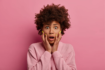 Portrait of shocked terrified woman grabs face, opens mouth as hears stunning news, has Afro...