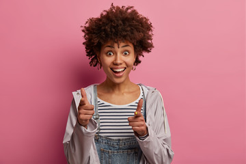 Check it out. Positive dark skinned woman points at you, introduces product, chooses someone, dressed in casual outfit, recommends something, smiles broadly, isolated on pink pastel background