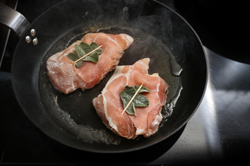 Chicken saltimbocca wrapped with air-dried ham and sage leaves are roasted in a black pan on the stove, copy space