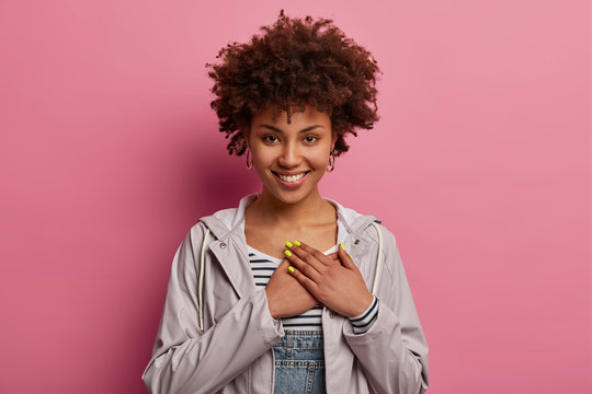Friendly positive curly woman presses hands to heart, smiles happily, expresses gratitude, thankful for heartwarming words, wears casual anorak, isolated on pink background, looks with admiration