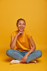 Vertical photo of good looking cheerful dark skinned lady keeps hand under chin, looks aside with broad smile, sits crossed legs on floor against yellow background, enjoys spare time with friends
