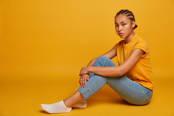 Photo of calm relaxed woman sits sideways against yellow background, has cornrows braids, wears casual t shirt and jeans, looks seriously at camera, feels relaxed. Copy space for your advertisement