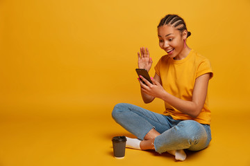 Horizontal view of ethnic millennial girl sits crossed legs on floor, enjoys browsing internet, has video call and waves palm, has online conversation, drinks coffee to go, isolated on yellow wall