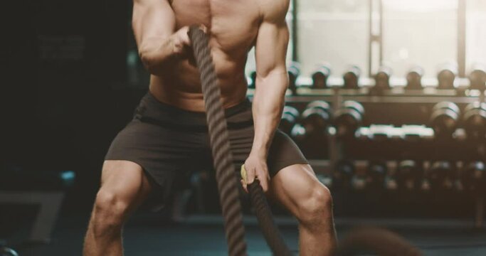 Attractive muscular man working out in the gym using battle ropes with high intensity, determined fitness man achieving his goals