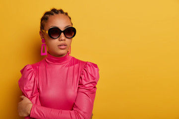 Portrait of stylish dark skinned woman has cornrow hairstyle, wears big shades and pink fashionable...