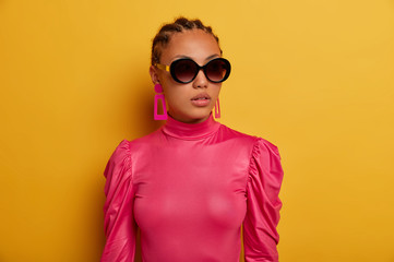 Stylish dark skinned woman wears stylish pink outfit, sunglasses and earrings, buys clothes of last...