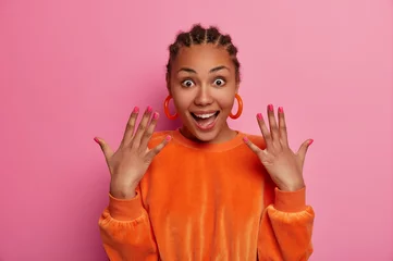 Poster Happy optimistic young woman raises hands, shows manicure, wears orange earrings, velvet jumper, has braided hairstyle, poses against pink pastel background, has positive surprised expression © Wayhome Studio
