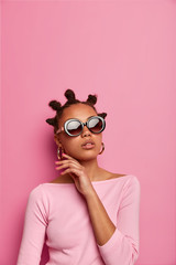 Vertical shot of charming tender woman touches face gently, has healthy smooth skin, elegant look, wears last fashion trendy sunglasses and pink jumper, has bun hairstyle. People, style, elegance