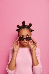 Beauty feminine fashion model wears stylish sunglasses, looks thoughtfully and with suspicion, has bun hairstyle, isolated on pastel rosy wall, empty space above for your promotion. Studio shot - obrazy, fototapety, plakaty