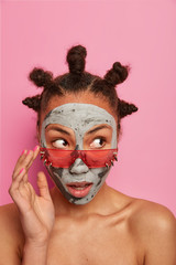 Portrait of surprised mixed race woman looks with wonder aside, wears trendy pink sunglasses, attends beauty spa, applies clay mask, cares about skin, has funny hair buns, stands naked indoor
