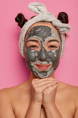 Close up portrait of pleased Asian woman applies clay mask, cleans skin from black dots, keeps hands under chin, wears headband, stands with bare shoulders against pink background. Spa and wellness