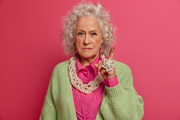 Attention to this information. Serious confident old woman raises finger, gives advice with convinced expression, warns you about danger, wears stylish clothes and accessories, stands indoor