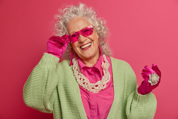 Cheerful curly haired senior woman with wrinkles and aging features keeps hand on rim of pink...
