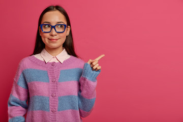 Beautiful clever female student in big glasses and jumper, introduces product promo, recommends click on link, shows item on pink background, presents new app for studying, shows best price. - obrazy, fototapety, plakaty