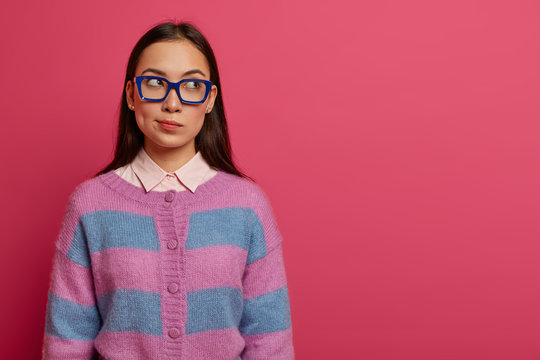 Horizontal shot of thoughtful brunette lady looks aside, thinks deeply about something, purses lips and has indecisive expression, wears spectacles, shirt and striped jumper, poses over rosy wall