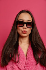 Studio shot of cheeky fashionable brunette lady has rounded lips, wears sunglasses and warm coat, has elegant look, going to have walk, poses against pink background. Fashion and style concept