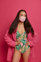 Serious thoughtful young woman thinks how to overcome virus, protects with medical mask, wears bathing suit and domestic robe, being ill, isolated on pink background. Health problems concept