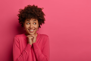 Portrait of positive dark skinned young woman keeps hands under chin, smiles toothily and looks aside, has pleased expression, wears crimson jumper, sees something appealing, creats plan in mind