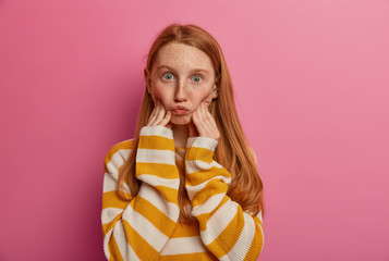 Photo of ginger freckled girl makes grimace, pouts lips, has widely opened eyes, wears loose jumper with stripes, poses indoor against pink background. Human face expressions, children concept