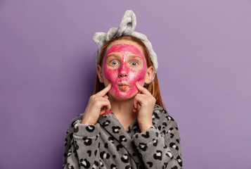 Photo of funny little girl keeps lips rounded, fingers on cheeks, applies face mask for reducing pimples, wears headband and soft robe, poses against purple background. Children, beauty concept