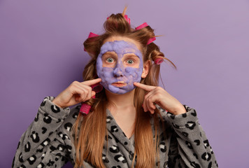 Photo of lovely freckled girl blows cheeks, makes funny face, wears hair curlers and cosmetic mask, prepares for special occasion, wants to look perfect, cares about skin and body, wears soft robe