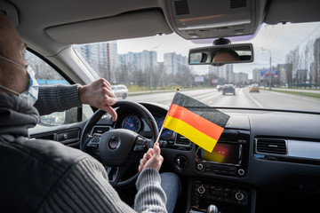 Middle aged man in the medical mask showing thumbs down with Germany flag driving a car. symbol of...