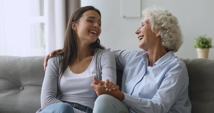 Loving middle aged mature smiling woman embracing young grown up daughter, chatting together in modern living room. Happy millennial girl talking bonding with older mother, sitting together on couch.