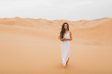 Fototapeta na wymiar Pretty portrait of young beautiful woman in sand dunes of moroccan Sahara desert. Brunette with long hair, eastern appearance. Bride in white silk dress.