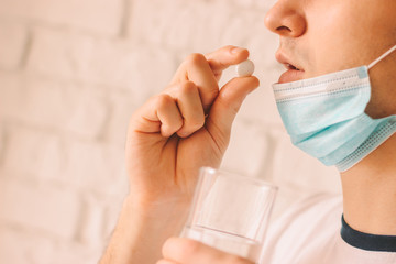 Closeup of young ill man in medical protective face mask taking white round pills with glass of...