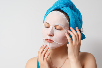 a young beautiful woman uses a moisturizing cosmetic fabric face mask with a towel wrapped around her head. on white background.