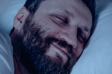 A lonely bearded man sleeps in bed at night and sees a good dream. Dreams, healthy sleep, smile, bedroom, pajamas, white bedding.