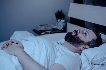 A lonely bearded man in bed at night cannot sleep, is unhappy and very tired. Sleep problems, stress, insomnia, bedroom, pajamas, white bedding.