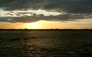 Pôr-do-sol em orla de rio com barcos em contraluz navegando e floresta na outra margem