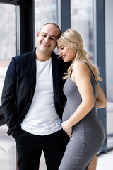 Fototapeta premium Young couple expecting baby standing together indoors near window. Young stylish family standing at home interior. Pragnancy concept.
