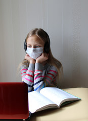 Blonde girl in a medical mask with headphones sits at a table with a computer and a book. Home schooling. The school is closed on quarantine due to the pandemic of Covid-19. Distance learning