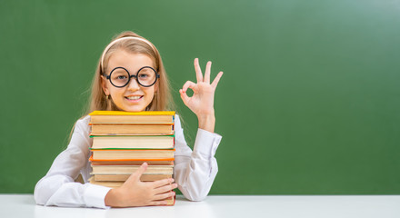 Smiling young girl wearing eyeglasses  sits with books near empty green chalkboard and shows okey gesture. Empty space for text © Ermolaev Alexandr