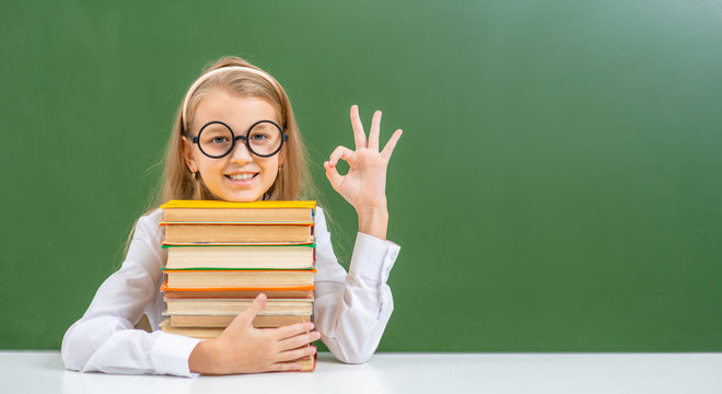 Smiling young girl wearing eyeglasses  sits with books near empty green chalkboard and shows okey gesture. Empty space for text