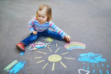 Fototapeta premium Adorable toddler girl drawing with colorful chalks on asphalt